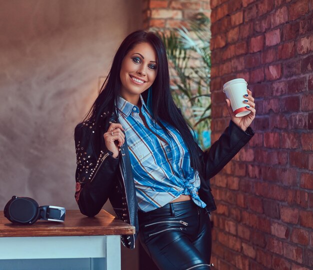 Retrato de una morena sensual sonriente posando en una elegante chaqueta de cuero y jeans apoyados en una mesa sostiene una taza de café en un estudio con un interior de loft.