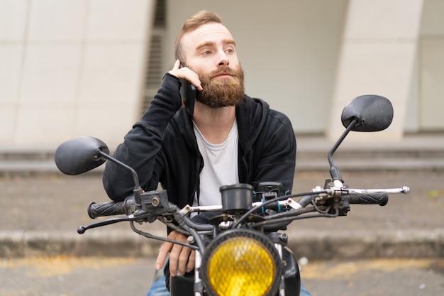 Retrato del motorista joven concentrado que habla en el teléfono móvil