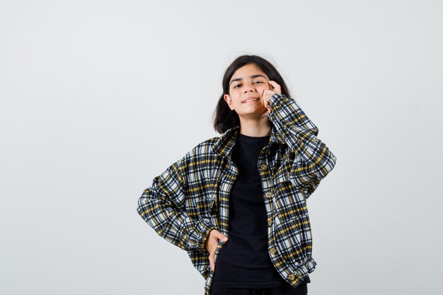 Retrato de muchacha adolescente sosteniendo el dedo en las sienes, manteniendo la mano en la cintura en camisa casual y mirando alegre vista frontal