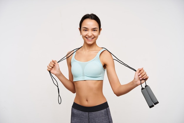 Retrato de muchacha asiática atractiva y deportiva con cabello largo oscuro. vistiendo ropa deportiva y sosteniendo una cuerda para saltar sobre su cuello. ten una sonrisa alegre. mirando a la cámara aislada sobre fondo blanco. Foto gratis