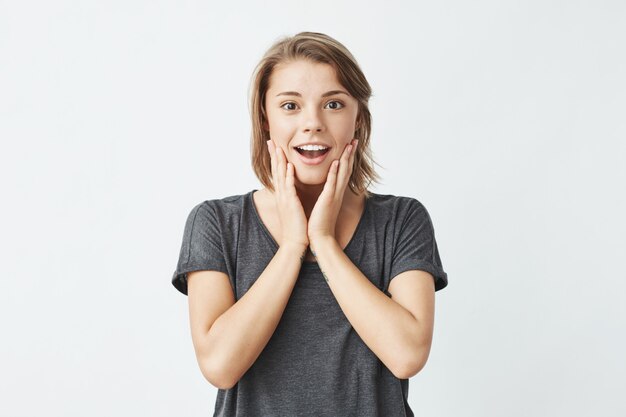 Retrato de la muchacha bonita joven sorprendida con la boca abierta que sonríe sosteniendo mejillas.