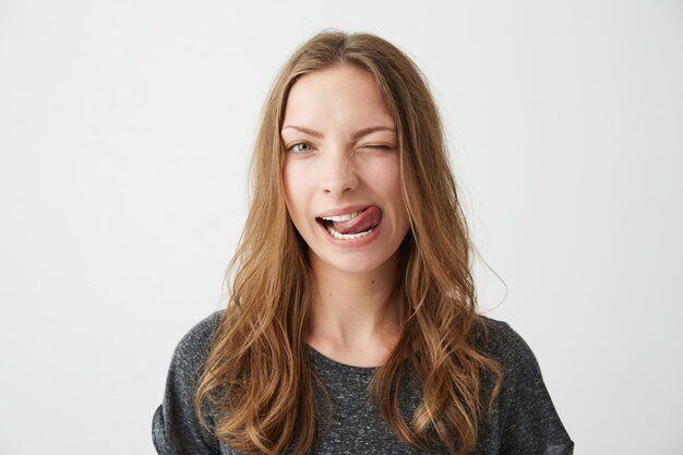 Retrato de la muchacha bonita positiva joven que guiña mostrando la lengua.