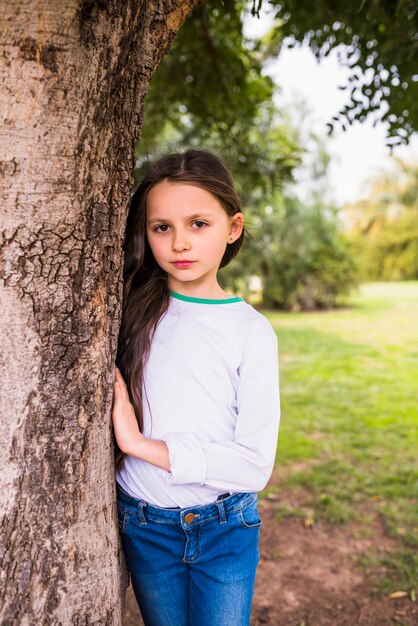 Retrato de una muchacha bonita que se coloca cerca de tronco de árbol en parque