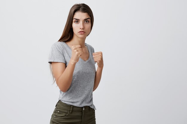 El retrato de la muchacha caucásica joven apuesta confiada con el pelo largo oscuro en camiseta gris y los vaqueros marrones que se colocan en lucha posan con la expresión seria y mala de la cara.