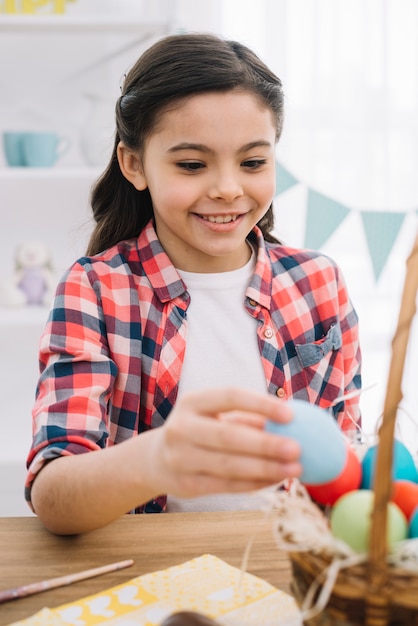 Retrato de una muchacha feliz que quita el huevo de Pascua azul de la cesta en la tabla