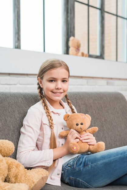 Retrato de una muchacha feliz que se sienta en el sofá gris que sostiene el oso de peluche que mira la cámara