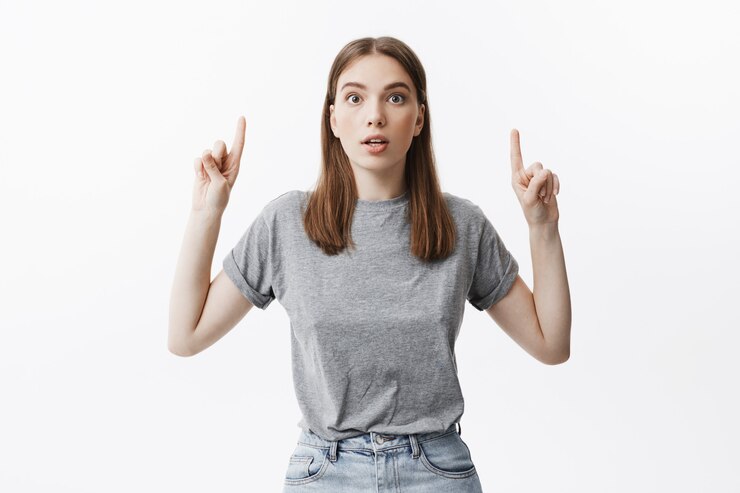Foto gratuito retrato de muchacha hermosa joven estudiante caucásico con cabello oscuro i ropa de moda casual con expresión sorprendida, apuntando al revés con ambas manos. copia espacio