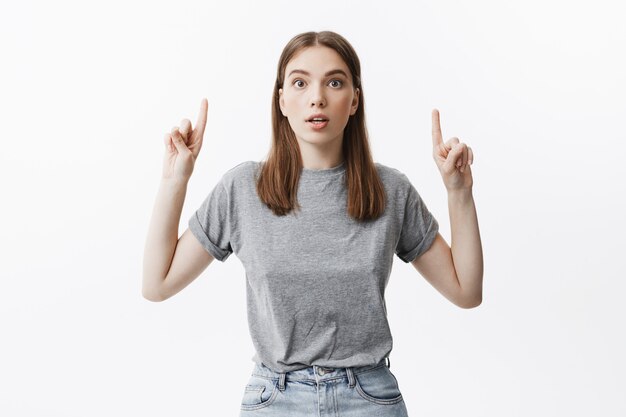 Retrato de muchacha hermosa joven estudiante caucásico con cabello oscuro i ropa de moda casual con expresión sorprendida, apuntando al revés con ambas manos. Copia espacio