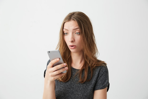 El retrato de la muchacha hermosa joven sorprendida que mira la pantalla del teléfono con la boca abierta que practica surf casó Internet.