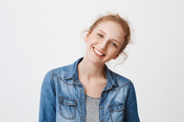 Retrato de muchacha pelirroja europea amigable positiva inclinando la cabeza hacia la derecha y sonriendo ampliamente, mirando a la cámara con ojos azules puros