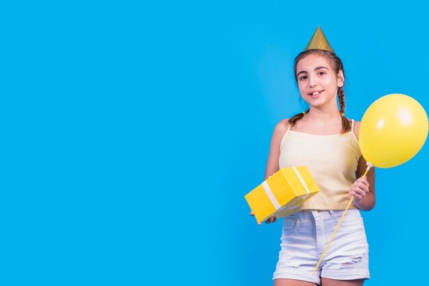 Retrato de una muchacha que sostiene la caja y los globos de regalo en su mano en superficie azul