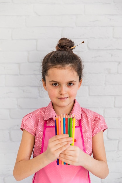 Retrato de una muchacha que sostiene los lápices multicolores en la mano que se opone a la pared de ladrillo blanca