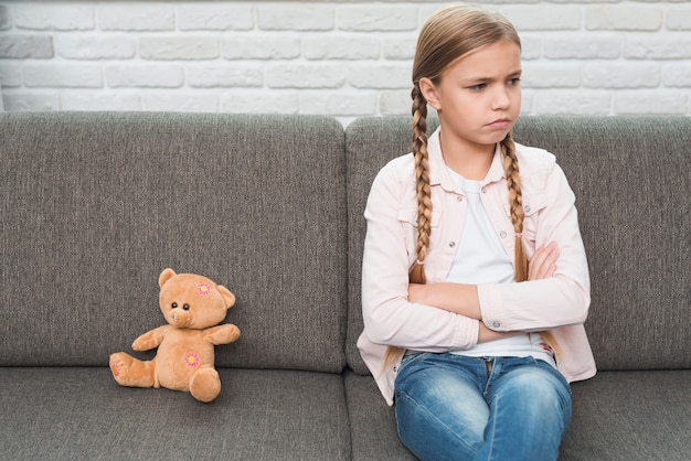 El retrato de una muchacha triste con los brazos cruzados que se sientan cerca del oso de peluche en el sofá gris