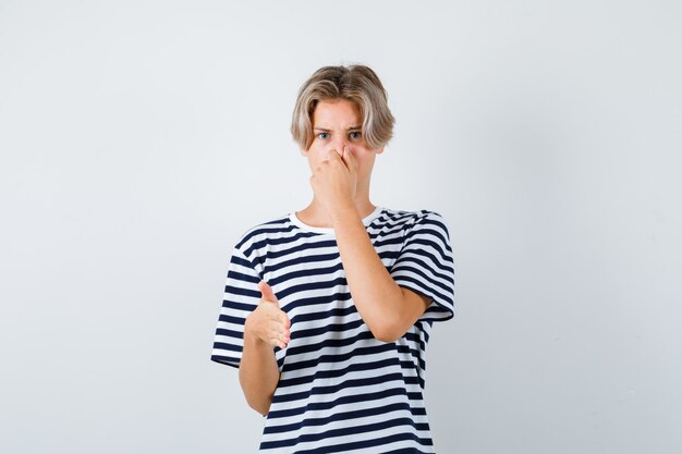 Retrato de un muchacho adolescente bonito que huele algo horrible, pellizca la nariz, que muestra la señal de pare en una camiseta a rayas y que mira la vista frontal disgustada