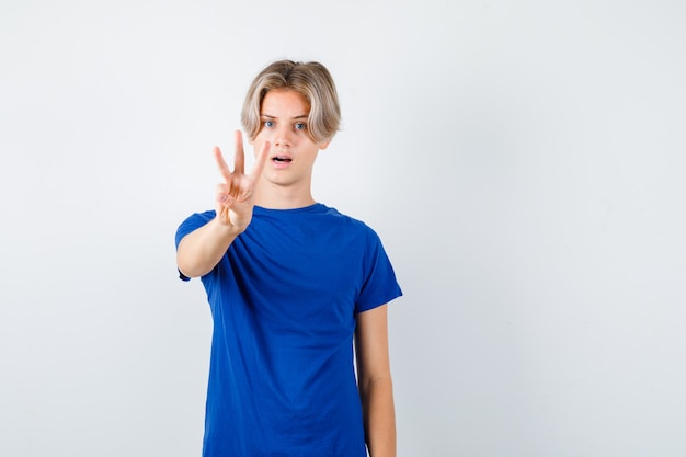 Retrato de muchacho adolescente guapo mostrando tres dedos en camiseta azul y mirando vacilante vista frontal