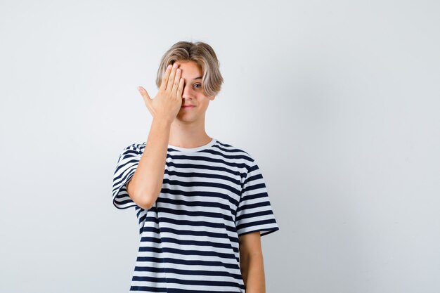 Retrato de muchacho adolescente que cubre su ojo con la mano en la camiseta y mira la vista frontal positiva