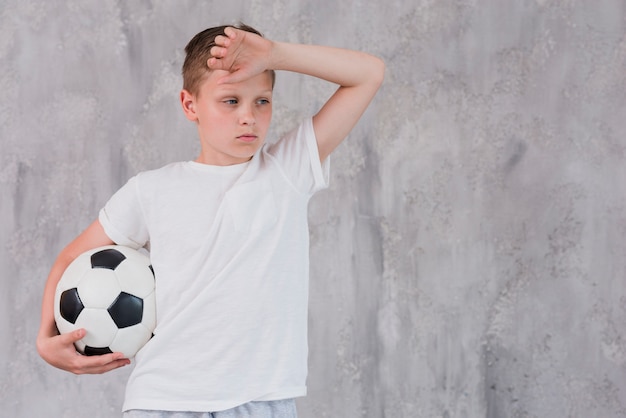 Retrato de un muchacho agotado que sostiene el balón de fútbol en la mano contra el muro de cemento