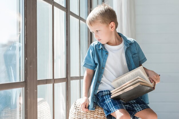 Retrato de un muchacho rubio que se sienta cerca de la ventana en luz del sol