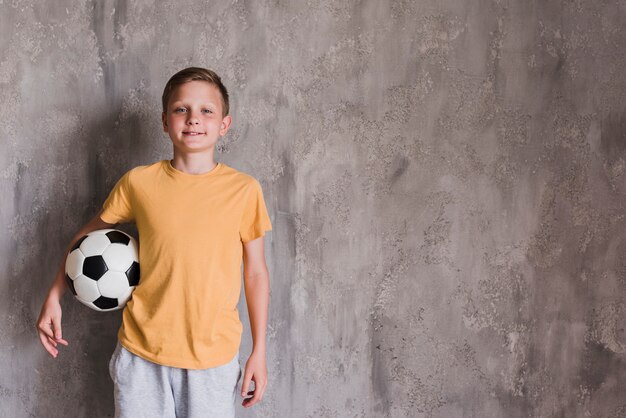 Retrato de un muchacho sonriente con el balón de fútbol que se coloca delante del muro de cemento