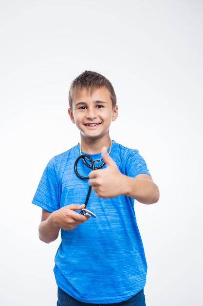 Retrato de un muchacho sonriente con el estetoscopio que gesticula los pulgares para arriba