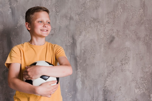 Retrato de un muchacho sonriente que sostiene el balón de fútbol delante del muro de cemento