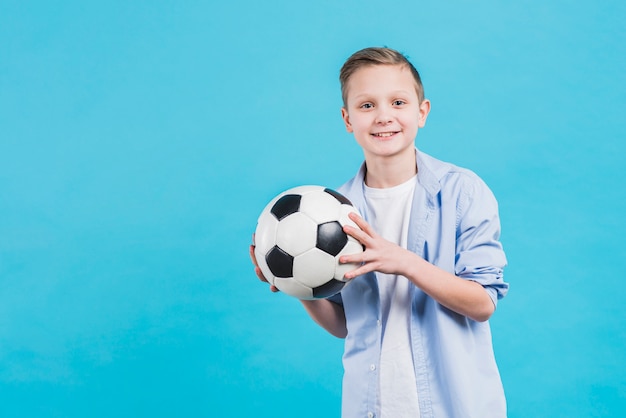 Foto gratuita retrato de un muchacho sonriente que sostiene el balón de fútbol en la mano que se opone al cielo azul