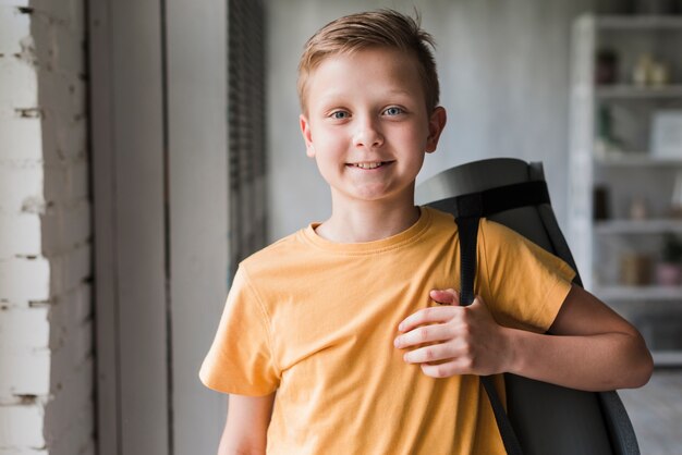 Retrato de un muchacho sonriente que sostiene la estera del ejercicio en su hombro