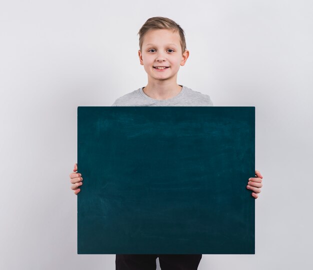 Retrato de un muchacho sonriente que sostiene la pizarra en blanco contra fondo gris