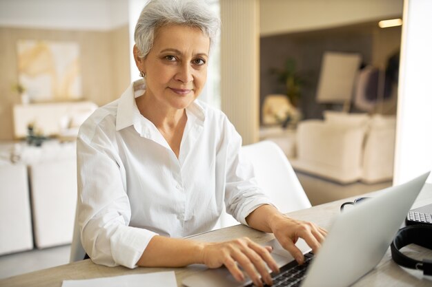 Retrato de mujer de 50 años de edad, alegre y de moda, escritora con camisa blanca, usando un dispositivo electrónico genérico para trabajar, escribiendo otro capítulo de su nuevo libro