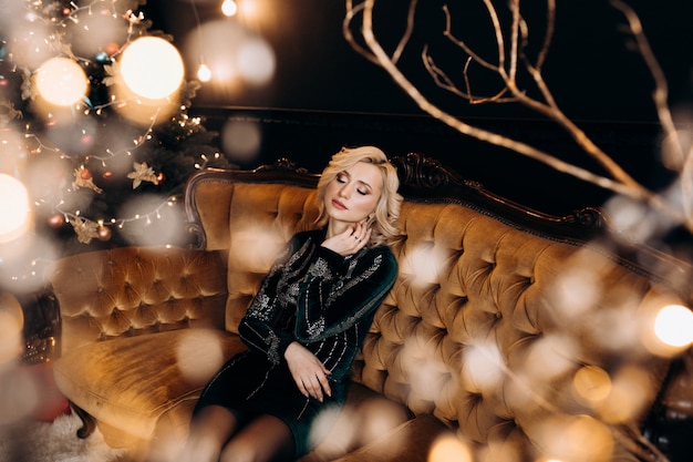 Retrato de una mujer adorable en vestido negro posando en un acogedor cuarto oscuro con decoración de Navidad