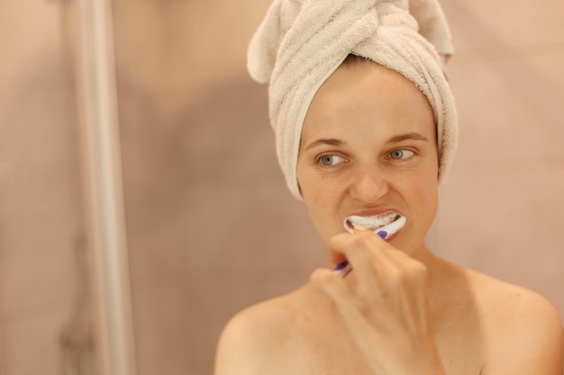 Retrato de mujer adulta joven atractiva con una piel perfecta con una toalla en la cabeza cepillándose los dientes, procedimientos de higiene en casa en el baño, sosteniendo el cepillo de dientes en las manos.
