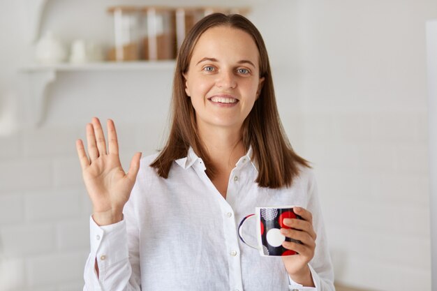 Retrato de mujer adulta joven feliz sonriente con camisa blanca mirando a cámara y agitando la mano, saludando, diciendo hola, expresando emociones positivas.