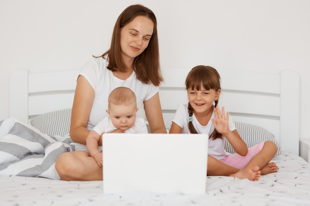 Retrato de mujer adulta joven independiente que trabaja en casa, mientras está sentado con sus hijos en la cama, autónomo y crianza de los hijos, familia posando en el dormitorio de luz.