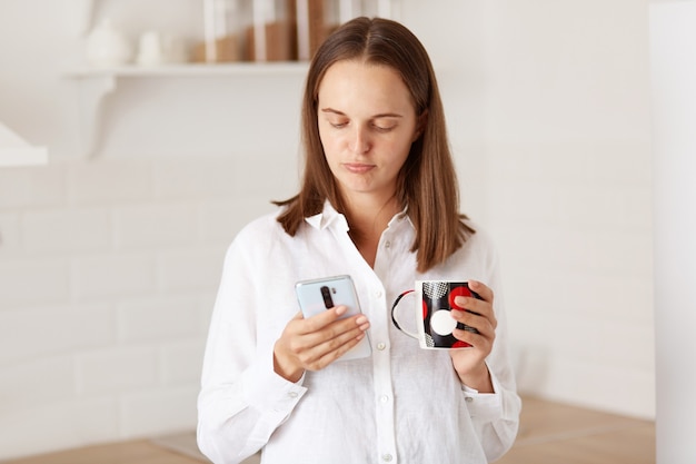 Retrato de mujer adulta joven infeliz de pie con el teléfono inteligente en las manos, tomando café o té en la cocina, expresando emociones negativas mientras lee el mensaje.