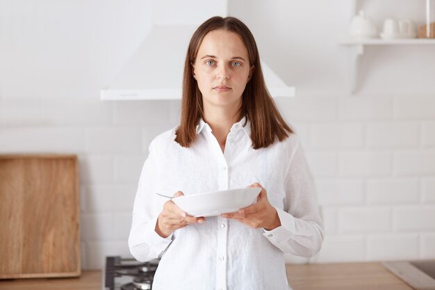 Retrato de mujer adulta joven molesta desayunando, sosteniendo en la cocina con el plato en las manos, mirando a la cámara con dolor, vistiendo ropa de estilo casual.