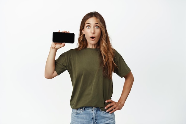 Retrato de una mujer adulta sorprendida que muestra una pantalla móvil con cara WOW, recomienda descargar la aplicación, demuestra el sitio web de la tienda, contra fondo blanco