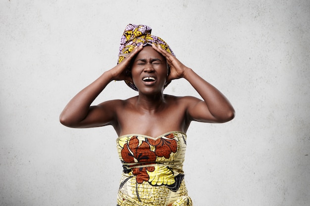 Retrato de mujer africana estresada con ropa colorida sosteniendo la cabeza con mirada dolorosa mientras sufre de migraña o dolor de cabeza.