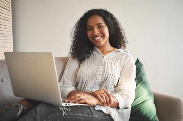 Retrato de mujer africana joven alegre en jeans y camisa sonriendo ampliamente mientras navega por internet en una computadora portátil genérica, disfrutando de una conexión inalámbrica de alta velocidad en la sala de estar