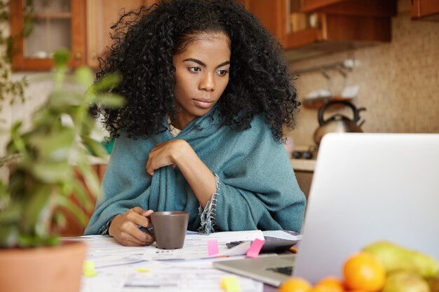 Retrato de mujer africana joven bebiendo té, mirando la pantalla del portátil con expresión enfocada