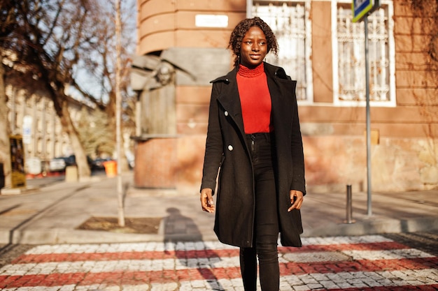 Retrato de una mujer africana de pelo rizado con abrigo negro de moda y cuello alto rojo caminando por el paso de cebra
