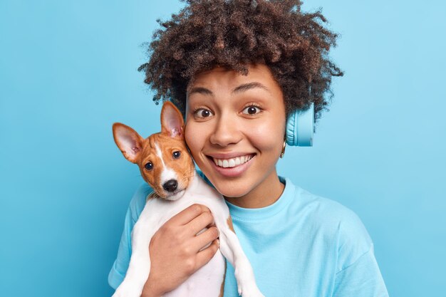 Retrato de mujer afroamericana alegre de buen aspecto sostiene un pequeño cachorro cerca de la cara sonríe agradablemente disfruta del tiempo libre con su perro favorito y usa auriculares estéreo aislados sobre una pared azul.