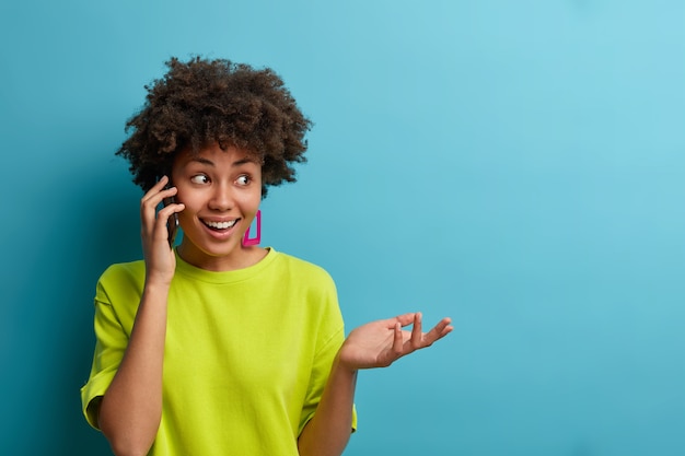 Retrato de mujer afroamericana alegre étnica pasa tiempo libre en casa chismeando, sostiene el teléfono inteligente cerca de la oreja, levanta la palma, vestida con camiseta casual, aislada en la pared azul, espacio en blanco