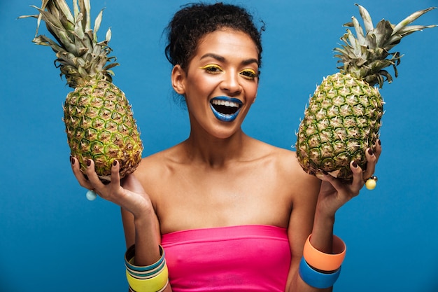 Retrato de mujer afroamericana alegre con maquillaje de moda sonriendo y sosteniendo dos piñas en ambas manos aisladas, sobre pared azul