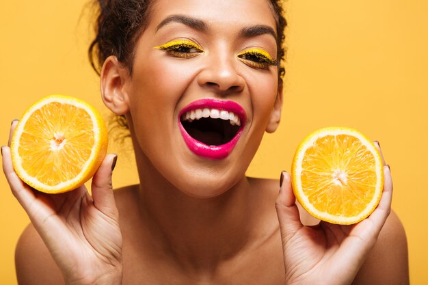 Retrato de mujer afroamericana atractiva con maquillaje de moda sosteniendo dos mitades de naranja en ambas manos aisladas, sobre pared amarilla