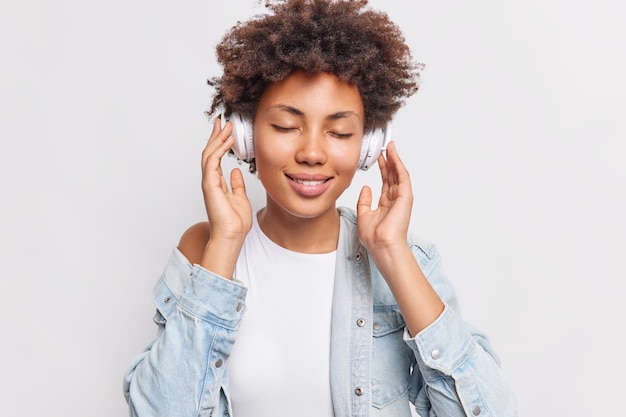 El retrato de una mujer afroamericana complacida disfruta de un buen sonido y su canción favorita mantiene las manos en los auriculares estéreo inalámbricos usa la mejor aplicación de música gratuita usa una camiseta blanca y una camisa de mezclilla posa en el interior