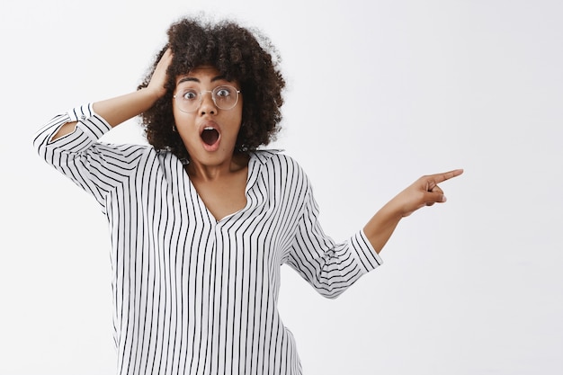 Retrato de mujer afroamericana confundida conmocionada y sin palabras con gafas y blusa a rayas apuntando hacia la derecha con la mano tirada sosteniendo el brazo en la cabeza dejando caer la mandíbula de tembló