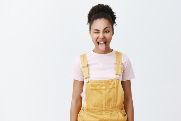 Retrato de mujer afroamericana feliz sin preocupaciones con un mono amarillo sobre la camiseta de moda, sacando la lengua y guiñando un ojo con alegría