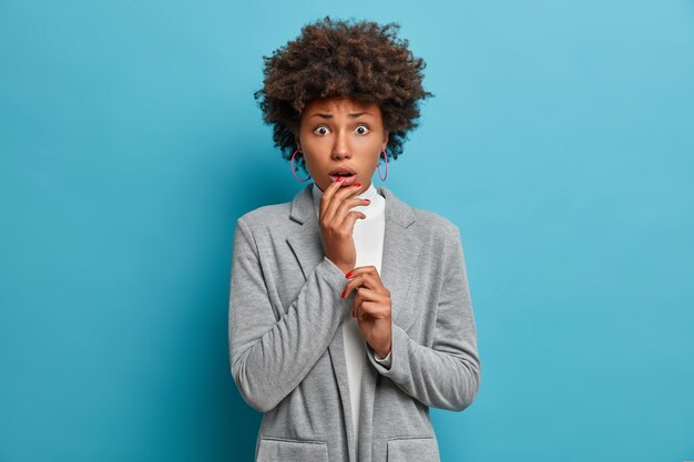 Retrato de mujer afroamericana joven preocupada preocupada mira con conmoción y pánico, sostiene la mano cerca de la boca abierta, ansiosa y asustada