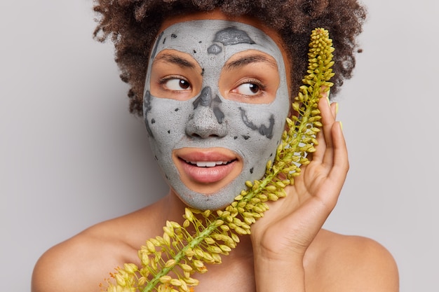 Retrato de mujer afroamericana pensativa aplica mascarilla de arcilla de hierbas nutritivas sostiene la planta