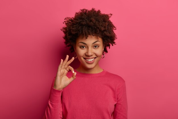 Retrato de mujer afroamericana satisfecha muestra buen gesto, dice excelente, anuncia buenas noticias, le gusta el producto y garantiza la mejor calidad, sonríe positivamente, da consejos y le gusta la elección